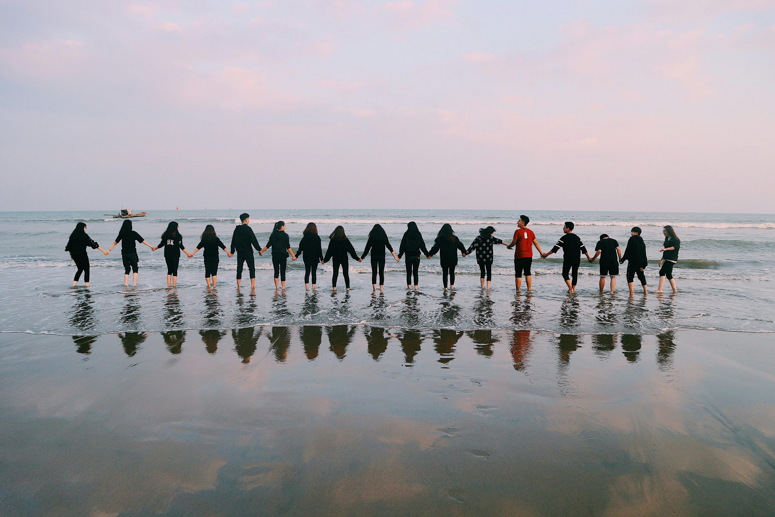 A large group holding hands at the beach during sunset, reflecting in the water.